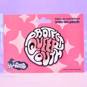 Op de afbeelding: Roze opstrijkbare patch met de woorden "PROTECT QUEER YOUTH" in een wit en roze ontwerp. De patch zit op een roze kaart met een sterrenpatroon en de tekst "Hella cute embroidered Iron-on patch".