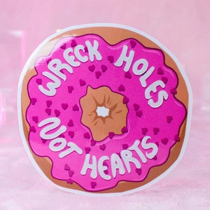 May include: A round sticker featuring a pink donut with brown frosting and a white center. The donut is decorated with small heart shapes and the words "WRECK HOLES NOT HEARTS" in white lettering.