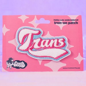 Op de afbeelding: Roze en blauwe geborduurde strijk-patch met het woord "Trans" in witte letters. De patch bevindt zich op een roze kaart met witte sterren en de tekst "Hella cute embroidered Iron-on patch".