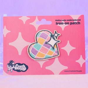 Op de afbeelding: Een roze en witte hartvormige strijkpatch met een geruit patroon in tinten roze, geel, groen en paars. De patch heeft een glinsterende witte ster bovenop en is verpakt op een roze kaart met witte sterren en de tekst "Hella cute embroidered iron-on patch".