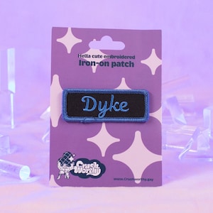 Può includere: Un toppa da stirare nero e blu con la parola "Dyke" ricamata in blu su uno sfondo nero. La toppa è su una carta viola con stelle bianche e il testo "Hella cute embroidered Iron-on patch".