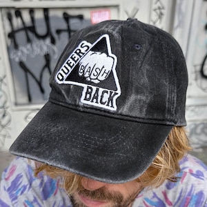 Könnte beinhalten: Schwarze Baseballkappe mit Used-Look. Auf dem weißen Aufnäher steht "QUEERS BASH BACK" in einem Dreieck. Die Kappe hat einen gebogenen Schirm und einen Stoffriemenverschluss.