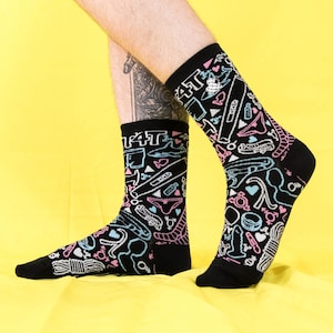 Puede incluir: Calcetines negros con un patrón repetido de varios juguetes sexuales en rosa, azul y blanco. La parte superior de los calcetines tiene la palabra "TAT" en blanco. Los calcetines están sobre un fondo amarillo.