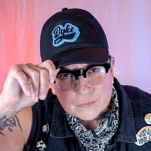 May include: A person wearing a black baseball cap with the word "Dyke" embroidered in blue. They are also wearing a black bandana, a black denim vest with various pins, and a pair of glasses. The person has tattoos on their arms.
