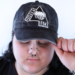 Peut inclure: Une casquette de baseball noire avec une finition délavée et usée. L'avant présente un écusson triangulaire blanc brodé contenant un graphique de poing blanc et le texte "QUEERS BASH BACK" en lettres majuscules blanches.