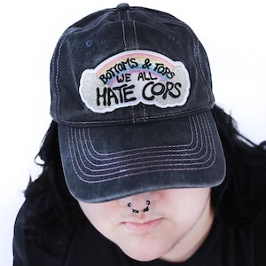 Könnte beinhalten: Eine schwarze Baseballkappe mit Used-Look und einem gestickten Aufnäher auf der Vorderseite. Der wolkenförmige Aufnäher zeigt einen Regenbogen und den schwarzen Sticktext: BOTTOMS & TOPS WE ALL HATE COPS. Die Kappe hat einen gebogenen Schirm.