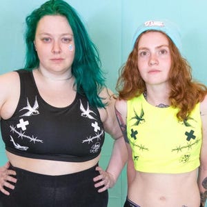 May include: Two people stand against a light blue backdrop, wearing crop tops and black trousers. One wears a black crop top with white bird and barbed wire designs. The other wears a neon yellow crop top with similar designs.