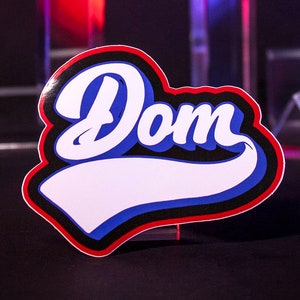 Könnte beinhalten: Aufkleber mit dem Namen "Dom" in einer weißen, blauen und roten Schriftart im Baseball-Stil. Der Aufkleber hat einen weißen Hintergrund mit blauem und rotem Umriss. Der Hintergrund ist schwarz mit Glasprismen.