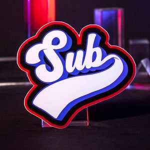 Könnte beinhalten: Ein Aufkleber mit dem Wort "Sub" in Weiß, Blau und Rot. Das Wort ist in einer stilisierten Schriftart mit schwarzer Umrandung gehalten. Der Aufkleber befindet sich auf einem transparenten Ständer, mit geometrischen Formen im Hintergrund.