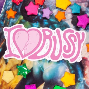 May include: A pink sticker with the text "I ❤️ BUSSY" in a bubble font, featuring a heart-shaped design with a stylized buttock illustration. The sticker is surrounded by colorful star-shaped origami.
