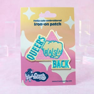 May include: Embroidered iron-on patch with the text "QUEERS BASH BACK" in teal and white on a pink triangle. The patch is on a light yellow backing with the text "Hella cute embroidered Iron-on patch".