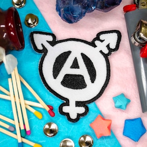 Peut inclure: Un écusson brodé blanc avec un symbole d'anarchie noir à l'intérieur d'un cercle, avec un symbole transgenre de chaque côté. L'écusson est sur une surface bleue avec une surface bleu clair à droite. Il y a des allumettes, un bocal en verre et un briquet sur la surface.