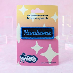 May include: A blue and black embroidered iron-on patch with the word "Handsome" in a cursive font. The patch is on a pink background with a white star design.