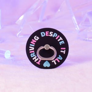 Könnte beinhalten: Ein schwarzer Telefonringhalter mit den Worten "THRIVING DESPITE IT ALL" in Pink, Blau und Weiß. Der Ringhalter hat einen silbernen Ring und ein kleines blaues Herz. Der Hintergrund ist ein weiches Lila.