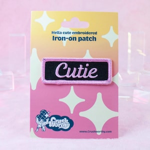Może przedstawiać: Różowo-czarna naszywka do prasowania z wyszytym różowym napisem "Cutie". Naszywka znajduje się na różowo-białej kartce z tekstem "Hella cute embroidered iron-on patch" i logo "Crush Worthy".