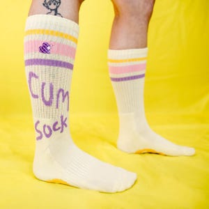 May include: Cream-colored knee-high socks with pink, purple, and yellow stripes. The socks have the words "CUM SOCK" in purple lettering. The background is a solid yellow.