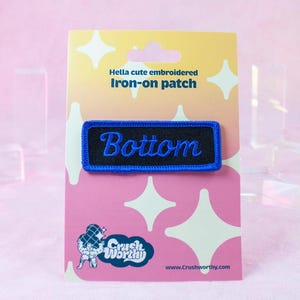 Pode incluir: Patch de ferro azul e preto com a palavra "Bottom" bordada em branco. O patch está em um fundo rosa e branco com estrelas e o texto "Hella cute embroidered iron-on patch".