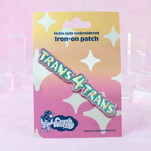 Op de afbeelding: Geborduurde opstrijkpatch met de tekst "TRANS 4 TRANS" in lichtblauwe en gele steken. De patch zit op een roze en gele kaart met de tekst "Hella cute embroidered Iron-on patch". De kaart heeft ook een logo en websiteadres.