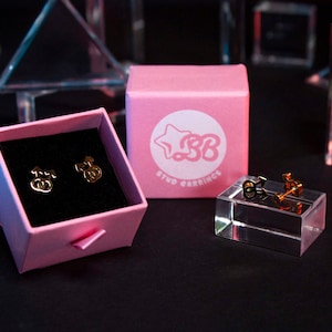 May include: Two gold stud earrings in a pink box with the text "Stud Earrings" and a star with the letters "SB". The earrings are on a clear display stand.