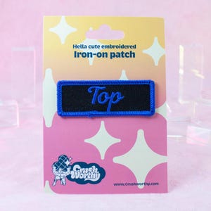 Op de afbeelding: Een blauwe en zwarte geborduurde strijk-op patch met het woord "Top" in witte letters. De patch is op een roze en witte achtergrond met de tekst "Hella cute embroidered iron-on patch" en het logo "Crush Worthy".