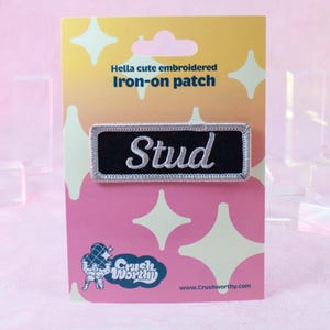 Op de afbeelding: Een zwart-witte strijkpatch met het woord "Stud" geborduurd in wit op een zwarte achtergrond. De patch bevindt zich op een roze en gele kaart met de tekst "Hella cute embroidered Iron-on patch" en het logo "Crush Worthy".