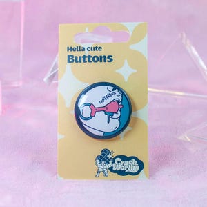 Könnte beinhalten: Ein runder Button mit einer Cartoon-Illustration einer Figur mit einem rosa Objekt im Mund, vor einem türkisblauen und schwarzen Hintergrund. Der Button befindet sich auf einer gelben Karte mit der Aufschrift "Hella cute Buttons" und einem Logo unten.