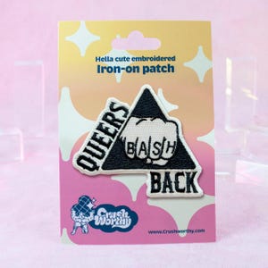 May include: Embroidered iron-on patch with the text "QUEERS BASH BACK" in black and white. The patch features a clenched fist inside a black triangle. The packaging reads "Hella cute embroidered Iron-on patch."