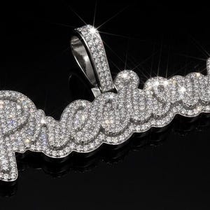 Puede incluir: Un colgante de plata con la palabra "Princess" escrita en cursiva y completamente incrustada de diamantes brillantes. El diseño del colgante es intrincado, con cada letra meticulosamente detallada. Sobre un fondo negro, los diamantes brillan.