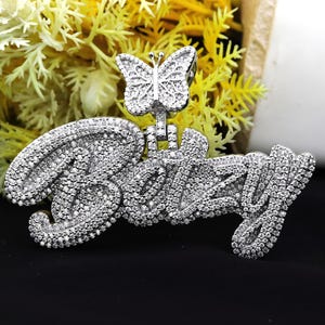 May include: A silver pendant with the name "Bizzy" spelled out in large, cursive letters. The pendant is covered in small, clear cubic zirconia stones. A small butterfly charm hangs from the top of the pendant.
