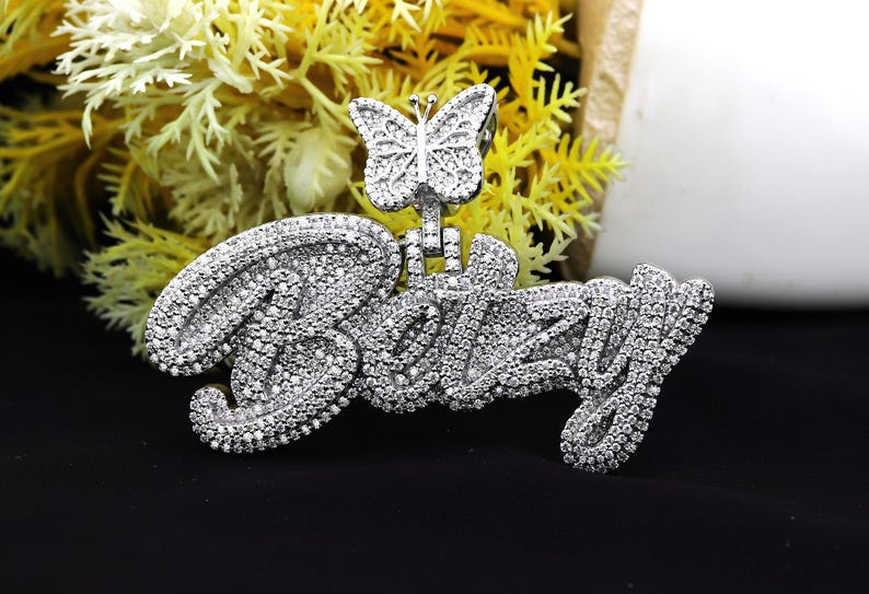 May include: A silver pendant with the name "Bizzy" spelled out in large, cursive letters. The pendant is covered in small, clear cubic zirconia stones. A small butterfly charm hangs from the top of the pendant.