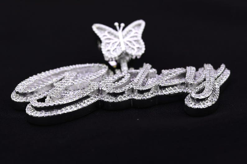May include: A silver pendant with the name "Izzy" spelled out in large, bold letters, encrusted with sparkling clear stones. A silver butterfly with clear stones is attached to the top of the pendant.
