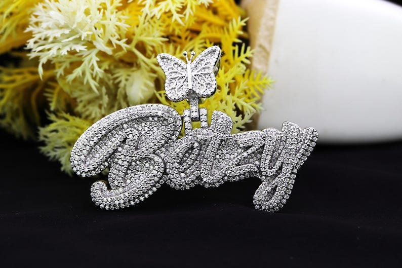 May include: A silver pendant with the name "Betsy" spelled out in sparkling rhinestones. A small butterfly charm with rhinestones is attached to the top of the pendant.