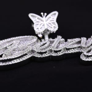 May include: A silver pendant with the name "Izzy" spelled out in large, bold letters, encrusted with sparkling white stones. A small butterfly charm with white stones hangs above the name.