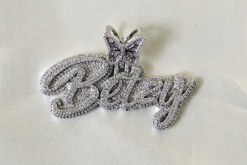 May include: A silver pendant with the name "Betsy" spelled out in sparkling cubic zirconia stones. A small butterfly charm with cubic zirconia stones is attached to the top of the pendant.