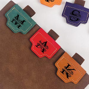 May include: A selection of colourful leather journal tabs, each with a different initial. The tabs are in orange, purple, red, green, and brown, and are attached to a brown journal. A red pen is also visible.