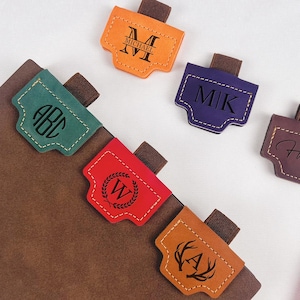 May include: A collection of personalized leather journal tabs in various colors, including orange, green, red, and brown. Each tab features a unique design with initials or names, perfect for organizing notebooks. A red pen rests nearby.