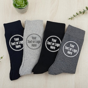 Op de afbeelding: Vier paar sokken met een witte cirkel op elke sok waarop "Your Text or Logo Here" staat. Twee paar zijn grijs, één is zwart en één is donkerblauw.