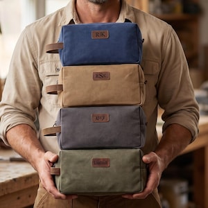 May include: A stack of four rectangular canvas toiletry bags in blue, tan, gray, and olive green. Each bag features a brown leather patch on the front with personalized lettering: R|K, RSK, W, and Lucas. Each bag includes a side leather handle.