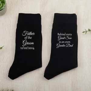 May include: Two black socks with white text. One sock says "Father of the Groom 12/02/2024". The other sock says "Behind every Great Son is an even Greater Dad".
