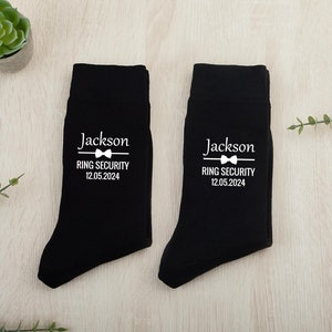 以下が含まれることがあります： 「Jackson Ring Security 12.05.2024」と蝶ネクタイのグラフィックが書かれた白いテキストがついた黒い靴下2足