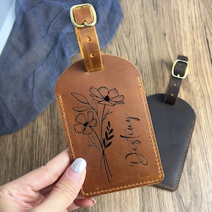 Puede incluir: Una etiqueta de equipaje de cuero marrón con un diseño floral y el nombre "Destiny" grabado en escritura cursiva. La etiqueta tiene una hebilla y una correa. Otra etiqueta marrón oscuro es visible en el fondo.