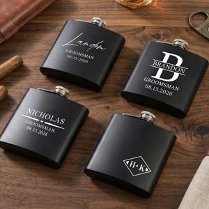 Pode incluir: Quatro frascos pretos com tampas prateadas, personalizados com nomes e datas em letras brancas. Um frasco tem o nome "Landon", outro "Brandon", um terceiro "Nicholas" e o último tem as iniciais "H.K.". Os frascos estão sobre uma superfície de madeira.