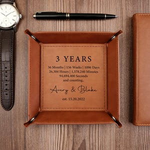 May include: A brown leather valet tray with the text "3 YEARS" and the names "Avery & Blake". The tray also includes the date "est. 15.20.2022" and time elapsed since the date. A watch, pen, and notebook are also in the image.
