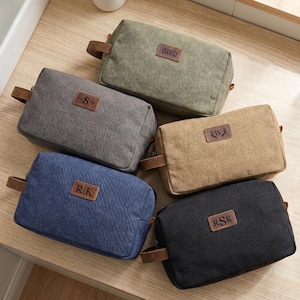 May include: Five rectangular toiletry bags in olive green, gray, tan, blue, and black. Each bag features a brown leather-like patch with personalized text including DAVID, S, W with antlers, R|K, and RSK, and a side handle for carrying.