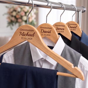 Può includere: Quattro grucce in legno chiaro con ganci in metallo argentato, ciascuna incisa con la parola Groomsman, un nome e la data 22.10.2024. Le grucce reggono abiti formali, tra cui una camicia bianca, un gilet grigio e pantaloni scuri.