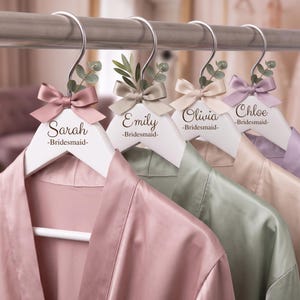 May include: Four white wooden hangers with silver hooks, each featuring a satin bow, a small sprig of greenery, and a name printed in script with the word Bridesmaid below. The names are Sarah, Emily, Olivia, and Chloe, each holding a satin robe.