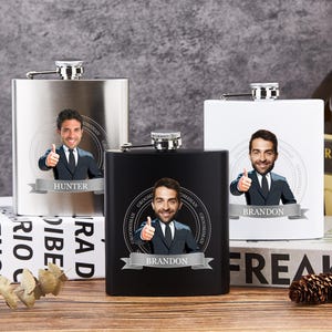 May include: Three hip flasks in stainless steel, black, and white, each with a personalized image and name. They have silver caps and name banners. The flasks are on a wooden surface, with books and a bottle of liquor in the background.