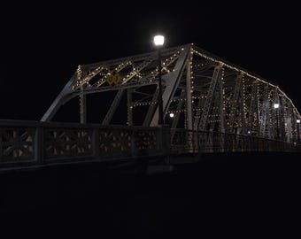 Può includere: Un ponte in metallo con luci a forma di ghirlanda. Il ponte è illuminato di notte e le luci si riflettono nell'acqua sottostante.