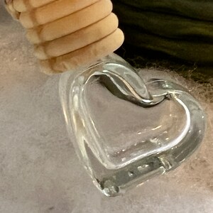 May include: A clear, heart-shaped glass pendant with a light-colored, cylindrical top. The pendant is suspended against a backdrop of white and green textures. The glass heart is the focal point, showcasing its transparent form.
