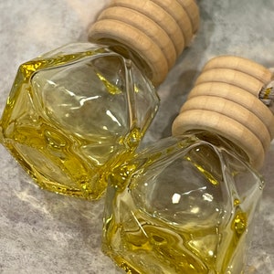 May include: Two clear glass car air fresheners with a geometric shape and a yellow tint. The fresheners are attached to wooden caps with a rope.